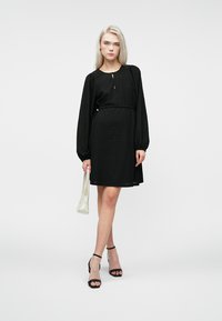 Μαύρο φόρεμα με μακριά μανίκια, υφή υφάσματος, ζώνη στη μέση, μέχρι το γόνατο. Συνοδευμένο από μια μικρή λευκή τσάντα και μαύρες σαγιονάρες.