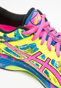 Botte de course jaune vif avec une tige texturée, ornée de motifs éclaboussés colorés, de lacets roses et d'une languette bleue avec un logo.