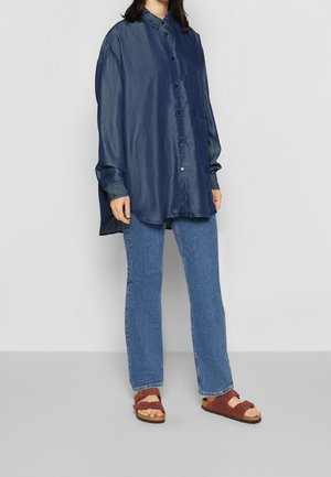 Person iført en oversized mørkeblå skjorte med knapper, lige blå jeans og brune åbne sandaler, der står mod en ensfarvet baggrund.