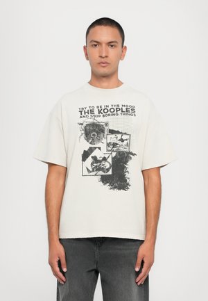 T-shirt bianco e morbido con stampa grafica nera, che presenta un collage di immagini e testo. Maniche corte e scollo rotondo.