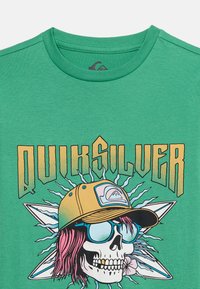 Πράσινο t-shirt με γραφικό ενός κρανίου που φορά γυαλιά ηλίου και κίτρινο καπέλο, ροζ μαλλιά, με το κείμενο "Quiksilver" από πάνω και στυλιζαρισμένες ακτίνες στο φόντο.