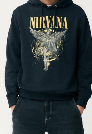 Black hoodie with yellow "NIRVANA" text above a distressed angel graphic, worn by a person with hands in dark jeans pockets.