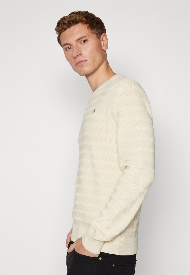 Kremowy sweter w paski z okrągłym dekoltem, z dżersejowymi mankietami i dołem wykonany z miękkiego materiału. Posiada małe logo na klatce piersiowej.