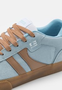 Light blue sneaker with brown leather accents, textured fabric upper, and brown laces. Features rubber sole and logo on side.
