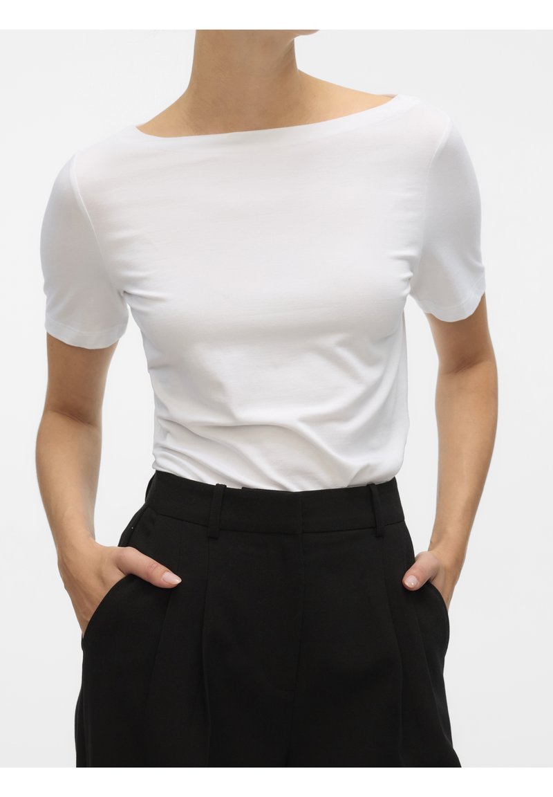 Femme portant un haut blanc ajusté à manches courtes, rentré dans un pantalon noir taille haute, les mains dans les poches, sur un fond uni.