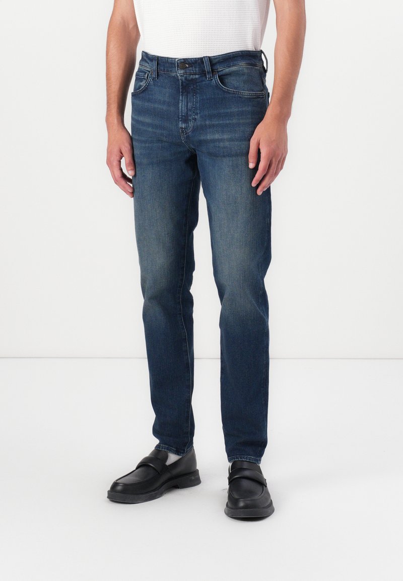 Donkerblauwe denim jeans met een rechte pijp, vijfzakkenontwerp en subtiele wassing. G gedragen met zwarte schoenen en een wit shirt.