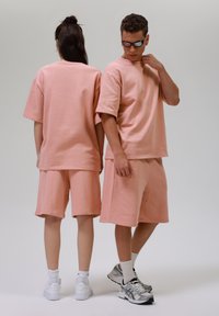 Dva modelky nosí odpovídající růžová oversized trička a šortky vyrobené z měkké tkaniny. Oblečení má volný střih a minimální detaily šití.