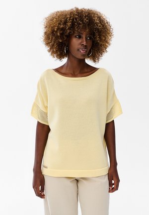 Kobieta o kręconych włosach, ubrana w żółtą, krótką sweterkową bluzkę i beżowe spodnie, stoi na białym tle, patrząc w lewo.
