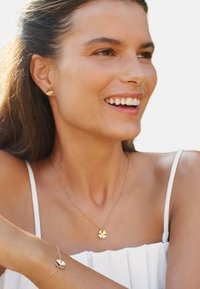 Guld smykkesæt bestående af et halskæde med en firkløver vedhæng, matchende armbånd og ørestikker. Modellen bærer en hvid top.