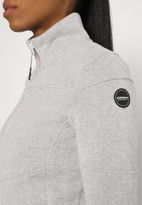 Grijze fleecejas met een hoge kraag, voorzien van een rits, structuurstof en een cirkelvormig logo op de linkerschouder.