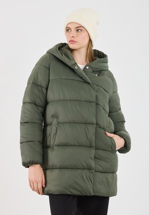 Jonge vrouw in een groene pufferjack en een crèmekleurige gebreide muts, die naar de zijkant kijkt met één hand in haar zak tegen een witte achtergrond.