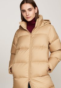 Jachetă puf bej cu glugă, cu închidere prin fermoar și linii de coasere vizibile. Textură netedă, mâneci lungi și buzunare laterale.