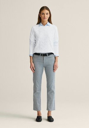 Kvinde stående iført en hvid tekstureret sweater over en blå skjorte med krave, grå stribede bukser, et sort bælte og sorte loafers.