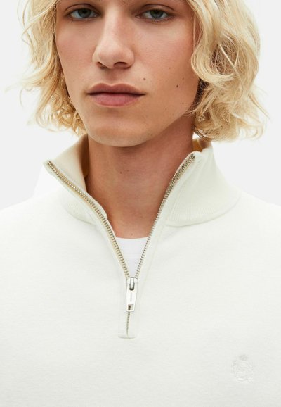 Weißer Reißverschluss-Pullover mit hohem Kragen, ausgestattet mit einem silbernen Reißverschluss und einem dezenten, gestickten Logo auf der Brust. Weiche Textur, minimalistisches Design.