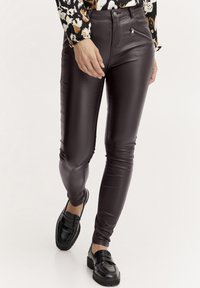Pantalon en simili cuir marron avec une coupe slim, présentant des détails de poche zippée et une texture lisse. Associé à des mocassins noirs.