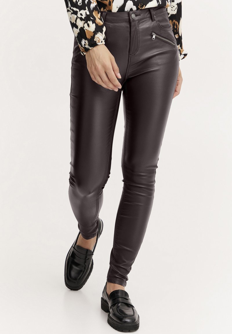Pantalon en simili cuir marron avec une coupe slim, présentant des détails de poche zippée et une texture lisse. Associé à des mocassins noirs.