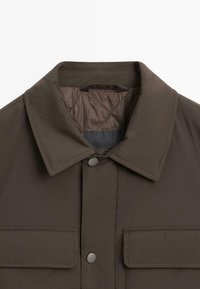 Brun vadderad jacka med klassisk krage, två framfickor, tryckknappar och en slät, matt yta. Fodret har quiltad textur.