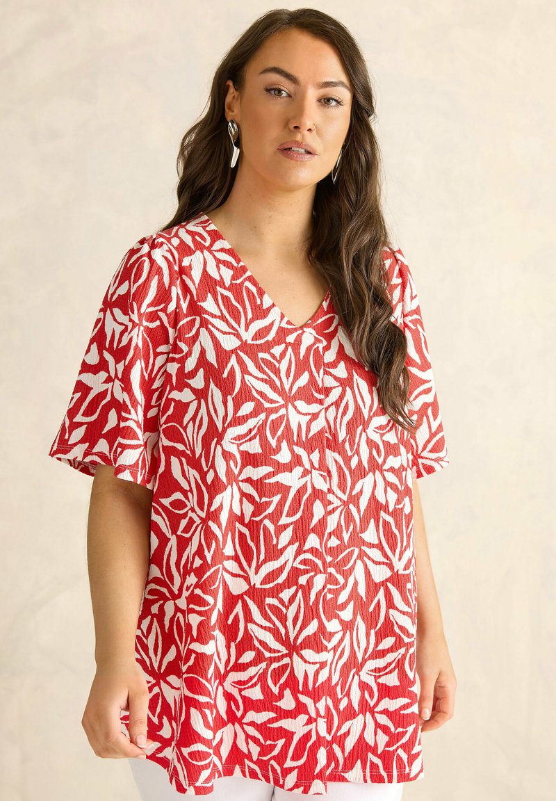 Mujer con cabello largo y castaño, vestida con una blusa de manga corta con escote en V y estampado de hojas rojas y blancas, y pendientes plateados, de pie frente a un fondo neutro.