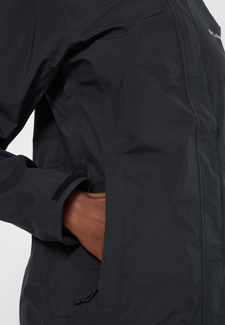 Main dans la poche latérale d'une veste imperméable noire avec un poignet réglable en Velcro.
