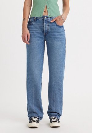 Person iført løstsiddende blå jeans, sort-hvide sneakers og en grøn ærmeløs knapbluse, der viser maven, står mod hvid baggrund.