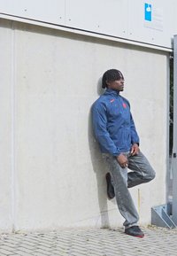 En person læner sig op ad en lys grå betonvæg, iført en blå Nike-jakke med et orange logo, slidte grå jeans og sorte sko.