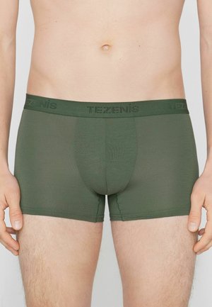 Männlicher Oberkörper, der olivgrüne Boxershorts mit einem markenprägten Taillenbund trägt, steht vor einem neutralen Hintergrund.