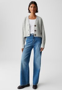 Donna che indossa un cardigan grigio chiaro con bottoni, una maglietta bianca, jeans blu a vita alta e larghi, e scarpe nere, in piedi davanti a uno sfondo neutro.