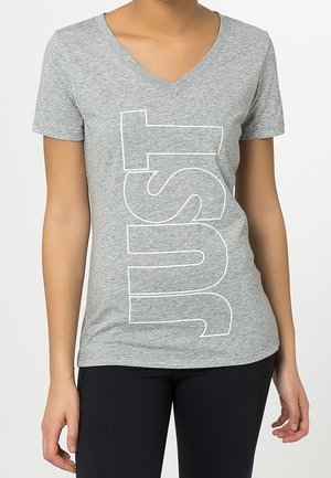 Kobieta w szarawym melanżowym T-shircie z dekoltem w serek z białymi obrysowanymi literami układającymi się pionowo w słowo "JUST", w połączeniu z czarnymi spodniami.