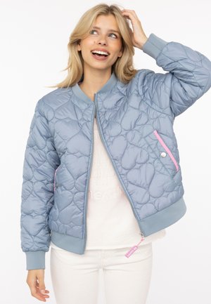 Junge Frau lächelt, trägt eine hellblaue gesteppte Jacke mit Herzmustern und rosa Reißverschlussdetails, weißes Hemd und Hose, Hand berührt Haare.