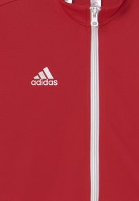 Röd jacka med hög krage, vit dragkedja och broderad Adidas-logotyp i vitt på vänster bröst. Materialet verkar slätt och sportigt.
