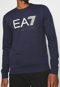 Mörkblå sweatshirt med långa ärmar, med en iögonfallande "EA7"-logotyp och texten "Emporio Armani" i kontrasterande vitt och svart. Material av bomullsblandning.