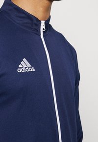 Marineblauwe ritsjack gemaakt van gladde stof, met een witte rits en het Adidas-logo in het wit op de linkerkant van de borst.