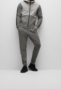Uomo che indossa un giubbotto con zip grigio BOSS e pantaloni jogger abbinati con scarpe da ginnastica nere, in piedi su uno sfondo grigio chiaro uniforme.