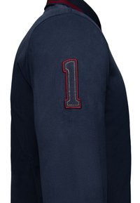 Marinblå polo t-shirt med en krage i röd och vit rand och ett stort broderat nummer "1" på vänster ärm. Mjuk, strukturerad bomullstyg.