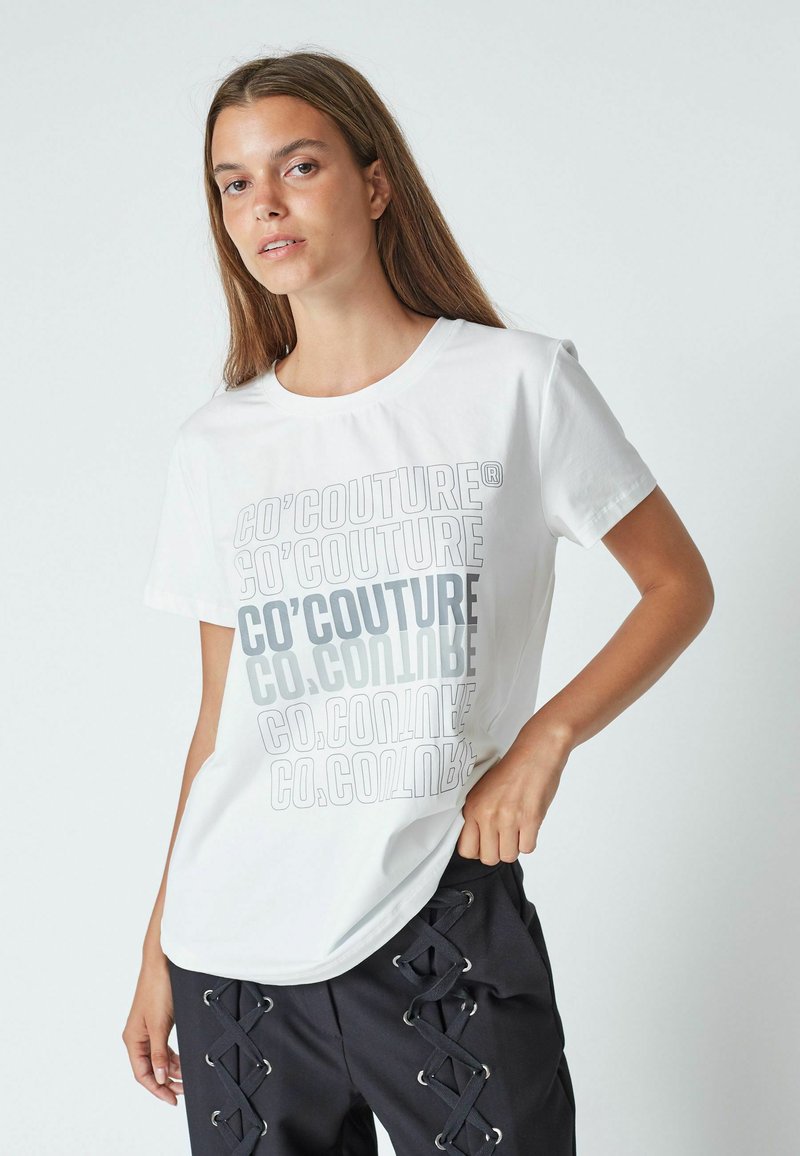 Jauna sieviete, tērpusies baltā T-kreklā ar atkārtotu "CO'COUTURE" tekstu un melnās biksēs ar krustveida šņorējumiem, stāv uz neitrāla fona.