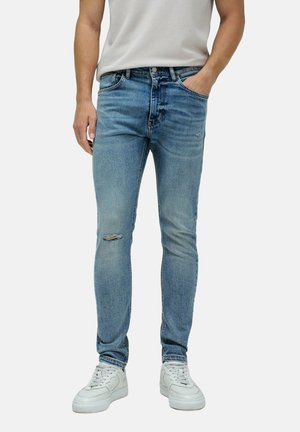 Vaqueros de mezclilla azul claro con corte ajustado, que presentan una rodilla desgastada, bolsillos estándar y cierre de botón. Combinados con zapatillas blancas.