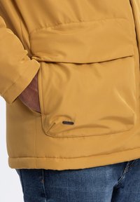 Gul puffjacka med en stor framficka, släta tyg och rundad nederkant. Har en liten logoplåt nära fickan.