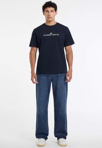 Tumesinine puuvillane T-särk, millel on valge "Guess Jeans" logo. Kombineeritud hästivoltitud siniste denimiste teksade ja valgete tossudega.