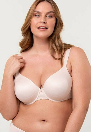 Kobieta o jasnej karnacji i długich jasnobrązowych włosach, ubrana w beżowy biustonosz, trzymająca jeden pasek, na tle jednolitego jasnego tła.