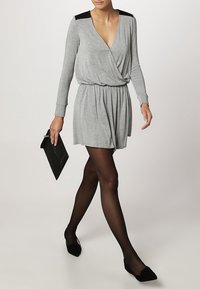 Robe enveloppante grise à manches longues avec des accents noirs aux épaules, taille élastique et jupe évasée. Portée avec une pochette noire et des collants opaques.