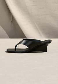 Sandale compensée noire à bout carré avec une large bride sur le pied, présentée sur un fond texturé neutre.