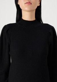 Pull en maille côtelée noire avec un col montant et des manches bouffantes structurées, présentant une texture douce et une silhouette ajustée.