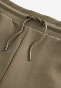 Shorts de sport vert olive avec cordon de serrage à embouts en métal et une finition en tissu doux et texturé. Pas de poches visibles.