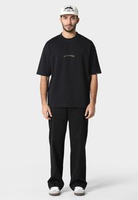 Black oversized t-shirt with "PAS DE MONACO" embroidered in gold, paired with black pants and white sneakers.
