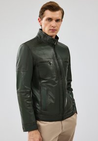 Giacca in pelle verde con colletto alto, chiusura con zip e due tasche sul petto. Presenta una texture liscia e dettagli di cucitura a contrasto.