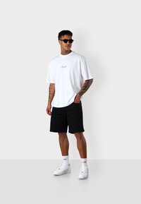 Weißes übergroßes T-Shirt mit besticktem Logo, schwarze Shorts, weiße Sneaker und Sonnenbrille. Vor einem neutralen Hintergrund präsentiert.