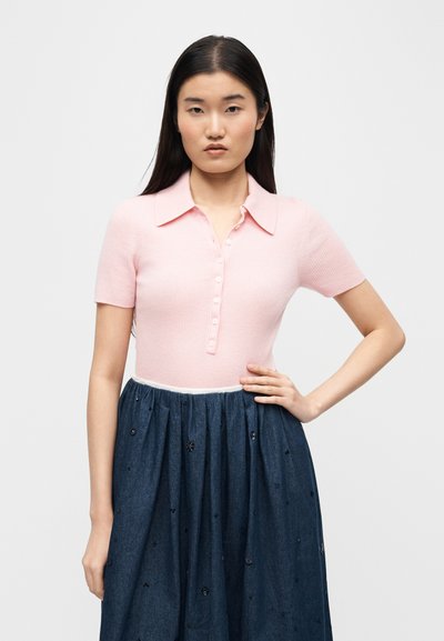 Νεαρή γυναίκα με μακριά σκούρα μαλλιά φοράει ανοιχτό ροζ κοντομάνικο μπλουζάκι με κουμπιά και σκουρόχρωμη μπλε φούστα με ψηλή μέση και διακοσμητικά στοιχεία.