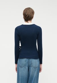 Tummansininen, pitkähihaiseksi muotoiltu paita, jossa on valkoinen kaulus ja sivusolmu yksityiskohtana, yhdistettynä korkeavyötäröisiin vaaleansinisiin farkkuihin.