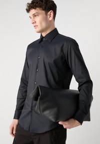 Camisa negra de manga larga con textura suave y cuello puntiagudo. El modelo sostiene un gran bolso de mano de cuero negro.