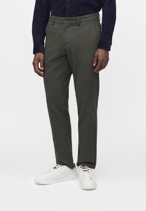 Mann, der dunkelgrüne Hose, weiße Sneakers und ein dunkles Langarmshirt trägt, steht vor einem hellen Hintergrund.